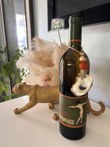 Dark green wine bottle with a green and orange label featuring a dancing figure, adorned with a string of beads and a seashell ornament on the neck.