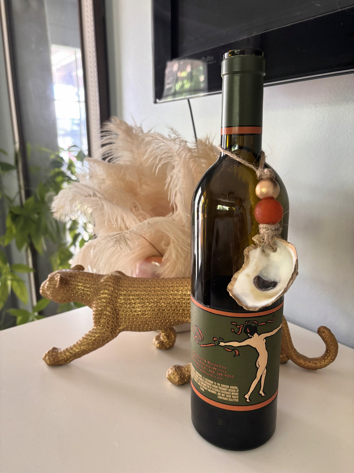 Dark green wine bottle with a green and orange label featuring a dancing figure, adorned with a string of beads and a seashell ornament on the neck.