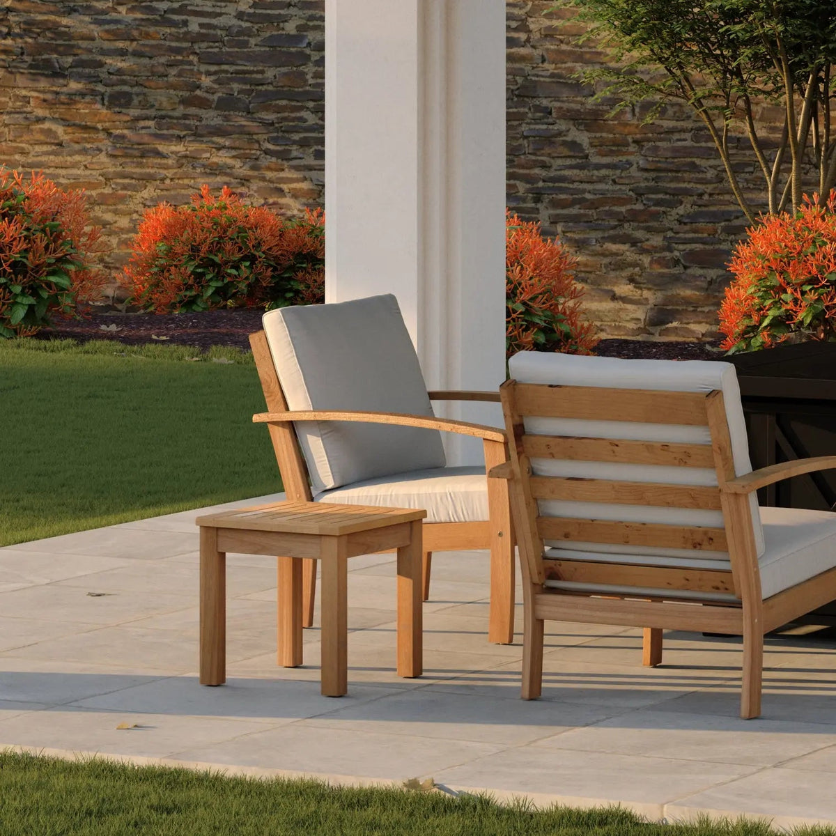 Wooden side table with slatted top and straight legs next to cushioned wooden armchairs on a stone patio with green lawn and stone wall backdrop