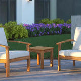 Small square wooden side table with straight legs positioned between two light wood armchairs with white cushions on a patio, with green grass and purple flowers in the background.