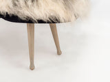 Two slender, rounded, light wood legs supporting a seat with fluffy, white and cream fur upholstery over a black base edge.