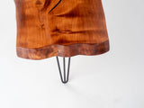 Close-up of a spalted maple wood table edge with rich brown tones and a natural wavy shape, supported by a slender black hairpin leg against a white background.