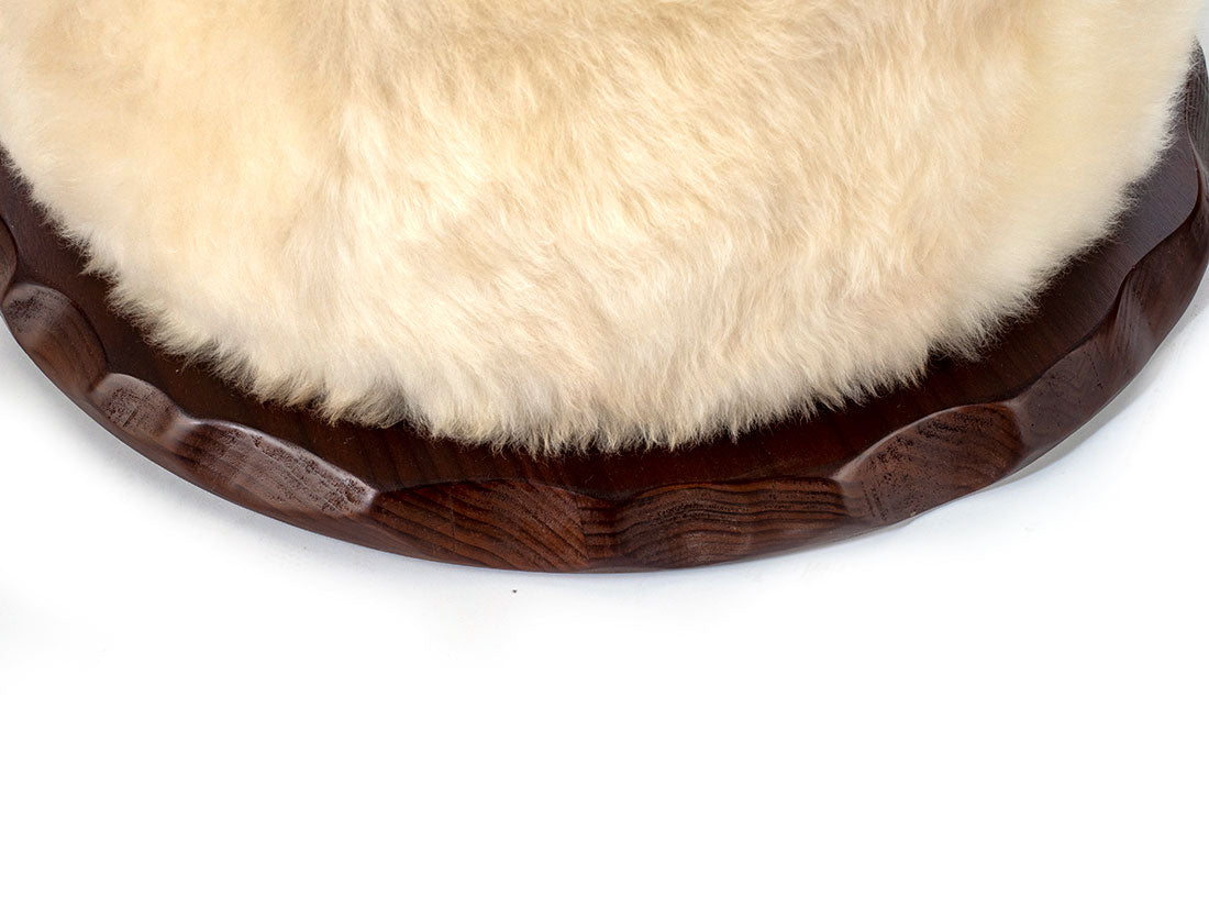 Close-up of round sheepskin cushion with fluffy cream-colored fur on carved dark wooden swivel base with scalloped edge detail.