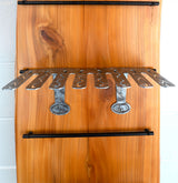 Vertical wine rack with stainless steel wine holder slots mounted on a smooth wooden board with visible grain and knots, framed by black metal rods at top and bottom.