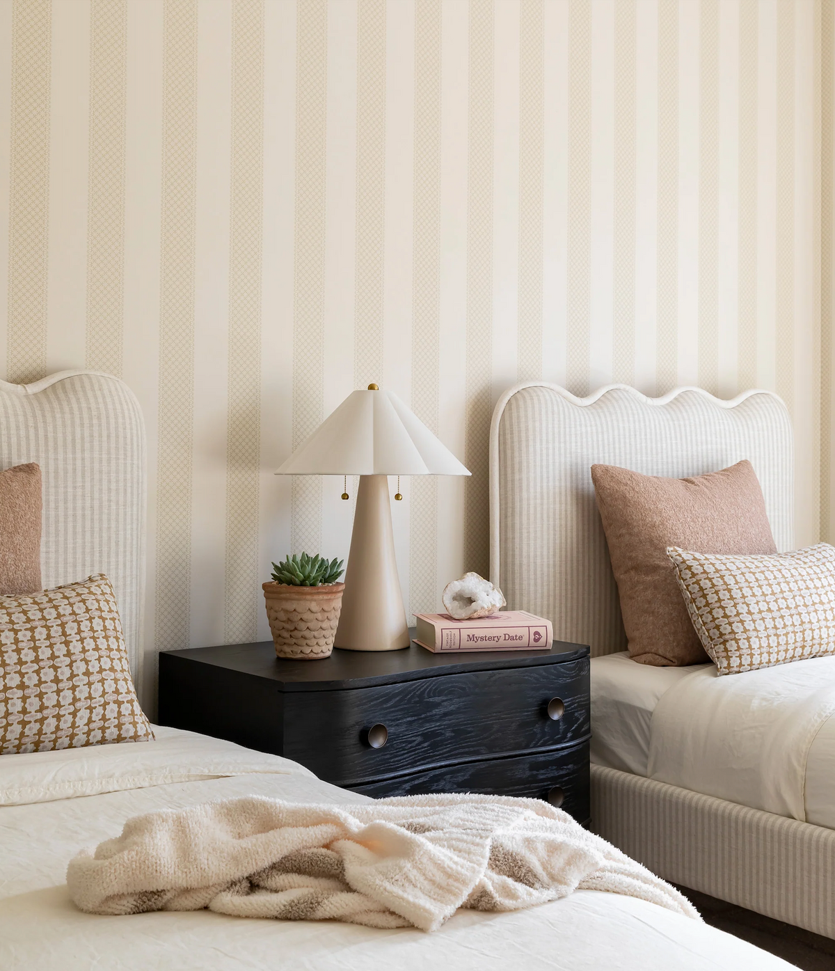 Two twin beds with scalloped beige headboards and patterned pillows flank a black nightstand holding a beige lamp, potted succulent, and pink book with flower decor.