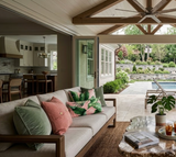 Beige cushioned outdoor sofa with green and pink palm leaf patterned pillows under a wooden vaulted ceiling opens to a patio with a pool and greenery beyond