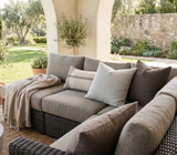 Outdoor woven sectional sofa with beige and brown cushions, featuring a chalk-colored striped pillow and a beige throw draped over one arm under a shaded archway.