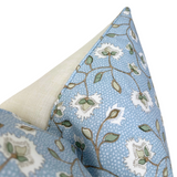 Corner detail of a pillow cover with beige fabric on one side and blue fabric patterned with white flowers and green leaves, featuring matching blue piping along the seam.
