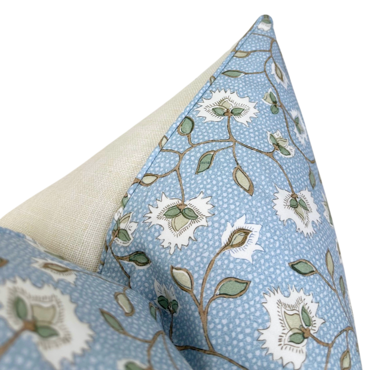 Corner detail of a pillow cover with beige fabric on one side and blue fabric patterned with white flowers and green leaves, featuring matching blue piping along the seam.