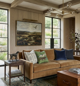 Brown suede sofa with four pillows in gray, green with horse print, and blue velvet, set in a room with large black-framed windows and wooden panel walls.