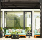 Outdoor seating area with two mint green cushioned wooden sofas featuring botanical patterned pillows and black side tables holding potted plants under a black pergola
