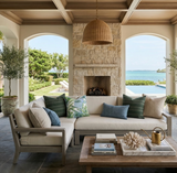 Beige cushioned outdoor sectional sofa with green striped and botanical patterned pillows in a covered patio with stone fireplace and ocean view through arches.