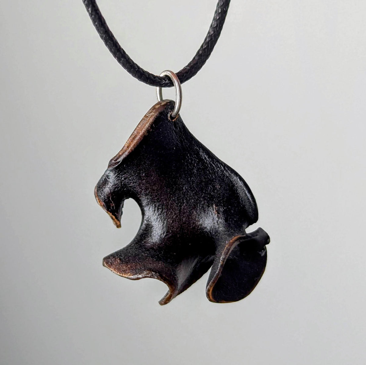 Black irregularly shaped pendant with smooth wave-like edges and subtle brown highlights, hanging on a black cord with a silver ring against a gray background.