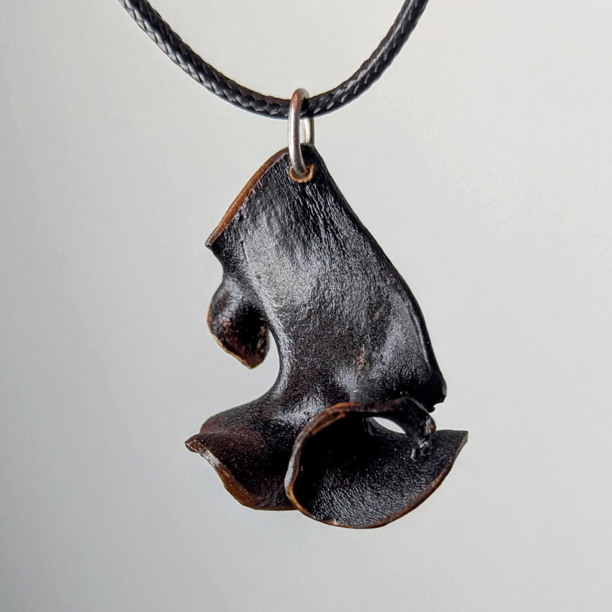 Curved, textured black pendant with organic, ruffled edges hanging from a braided black cord against a plain light background