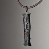 Dark textured cylindrical pendant resembling bark with two irregular translucent amber insets, hanging from a dark cord against a neutral background.