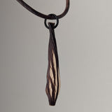 Elongated teardrop-shaped necklace pendant with dark brown textured outer edges and a central lighter section resembling wood grain on a dark brown cord.