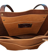 Interior view of a brown leather tote showing a large pocket with brass rivets and a dark brown leather label, with matching shoulder straps visible at the top.