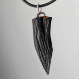 Close-up of a dark, textured wooden pendant shaped like a narrow, pointed leaf hanging from a black braided cord with a silver loop.
