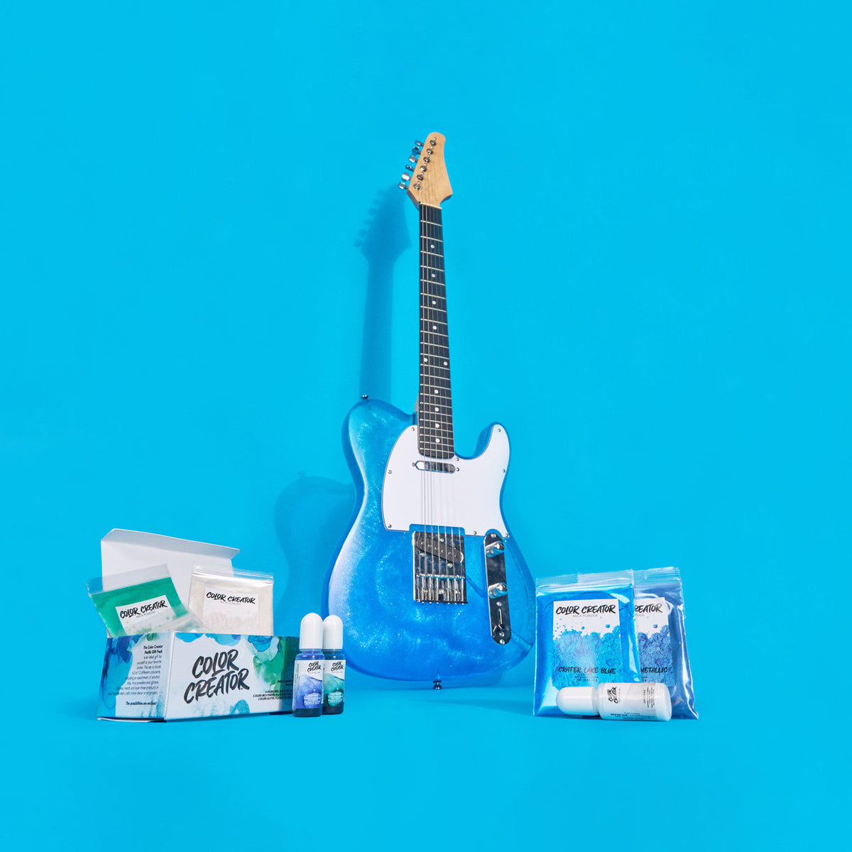 Electric guitar with a shimmering blue body and white pickguard, displayed against a bright blue background with color pigment packets and small bottles.