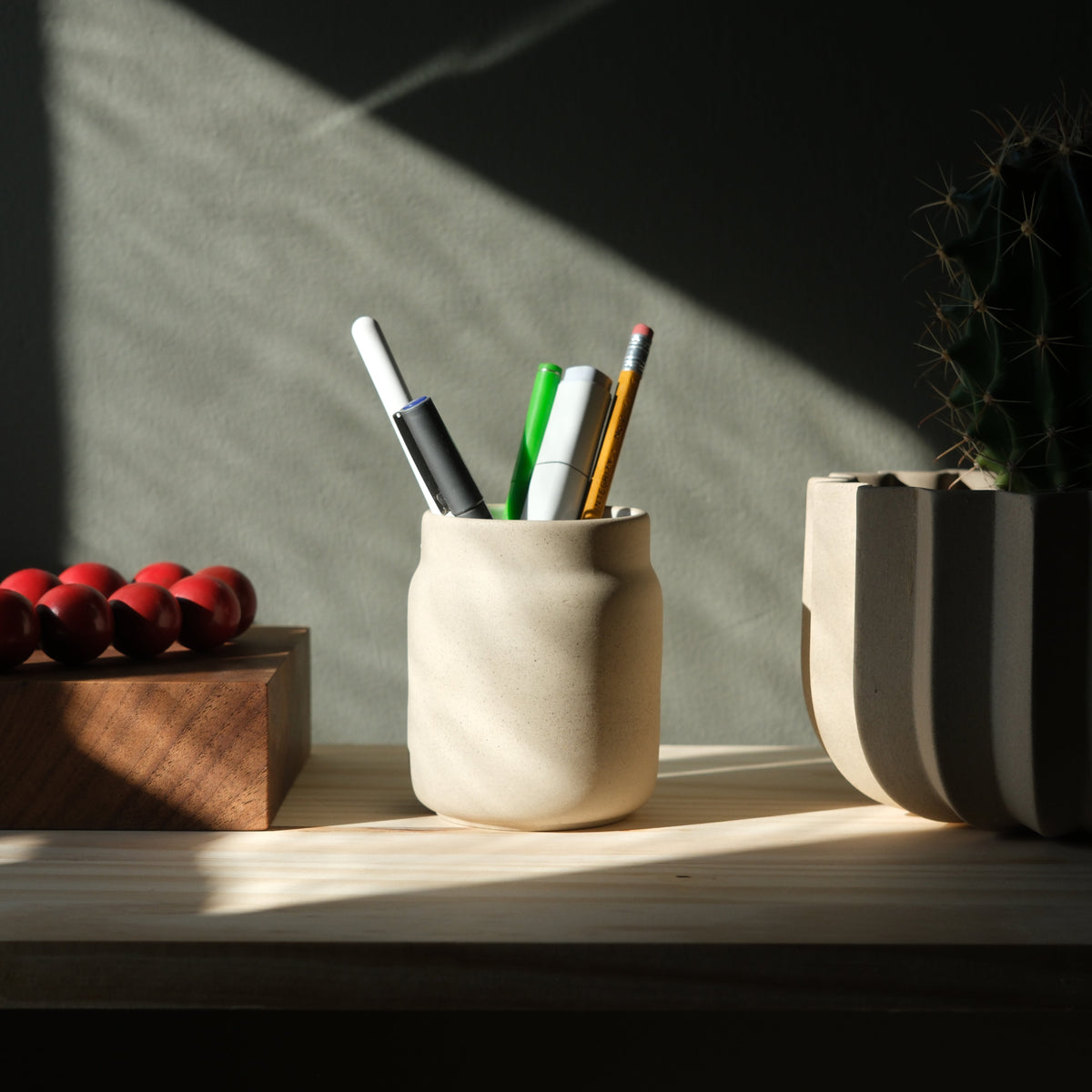 Matte beige ceramic pen holder containing pens and a pencil on a light wood surface, flanked by a red bead bracelet on a wooden block and a striped ceramic pot with a cactus, lit by sunlight.