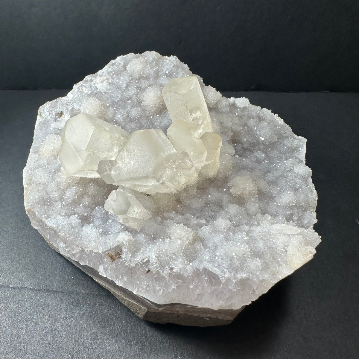 Cluster of translucent calcite crystals growing on a bed of white druzy quartz on an irregularly shaped agate base, displayed on a black surface.