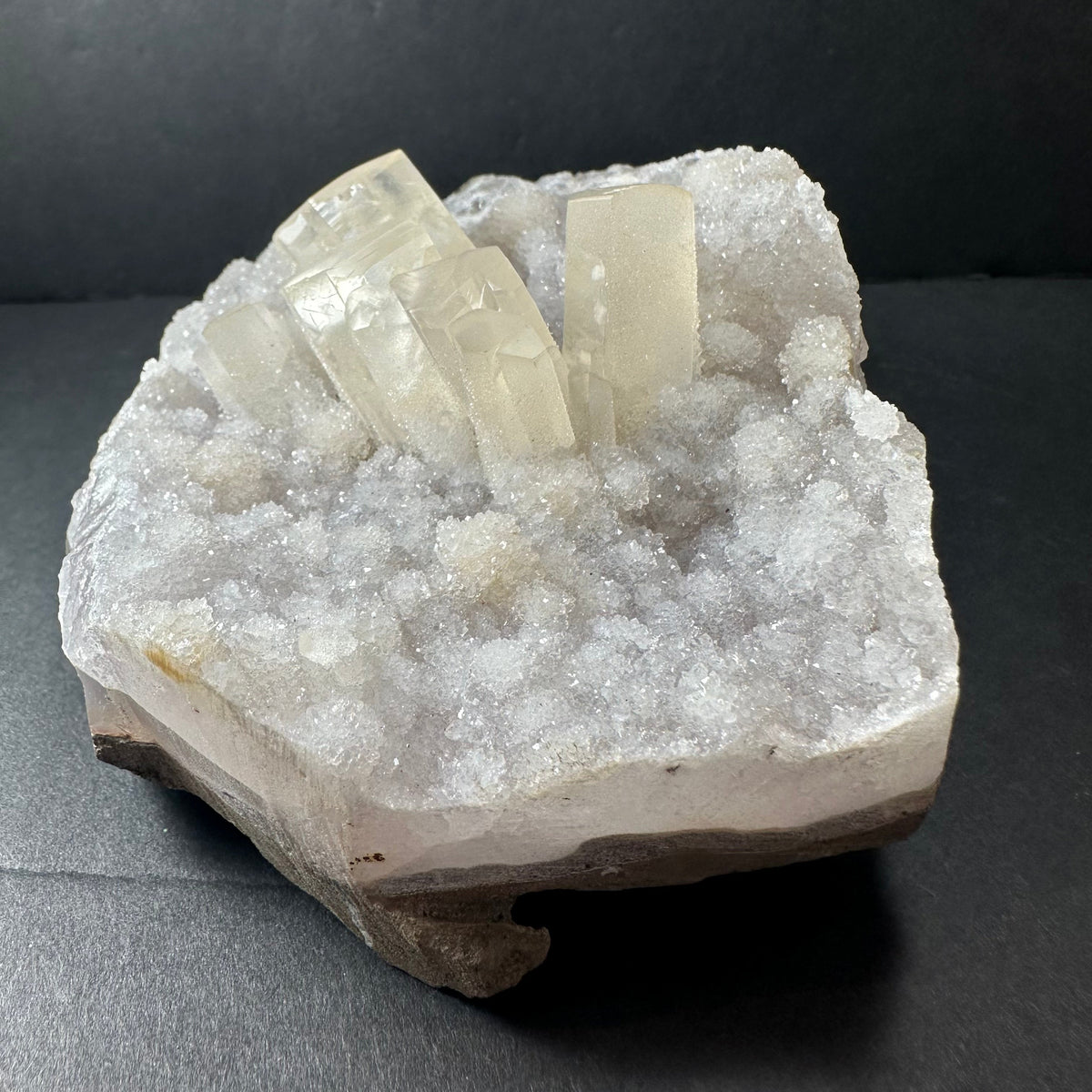 Clear druzy calcite crystal cluster with hexagonal prisms atop a sparkling white agate base with a rough bottom edge against a dark background