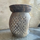 Carved soapstone essential oil burner with lattice pattern on curved body and dark marbled dish on top, set against a textured beige background.