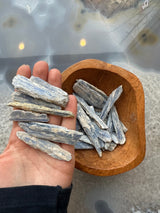 Hand holding several elongated raw blue-grey kyanite blades above a smooth wooden bowl filled with similar blades on a stone surface with light reflections.