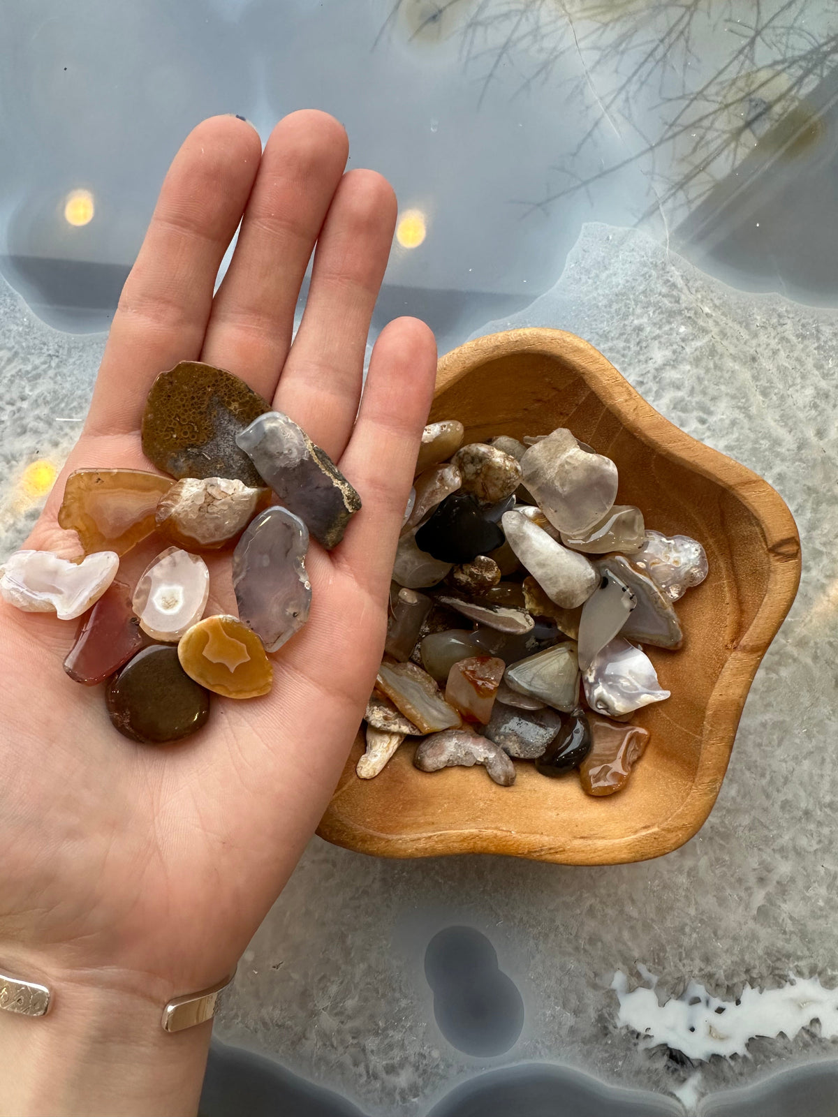 Hand holding various polished agate pieces in shades of brown, gray, and white, next to a wooden bowl filled with more similar agate stones on a reflective surface.