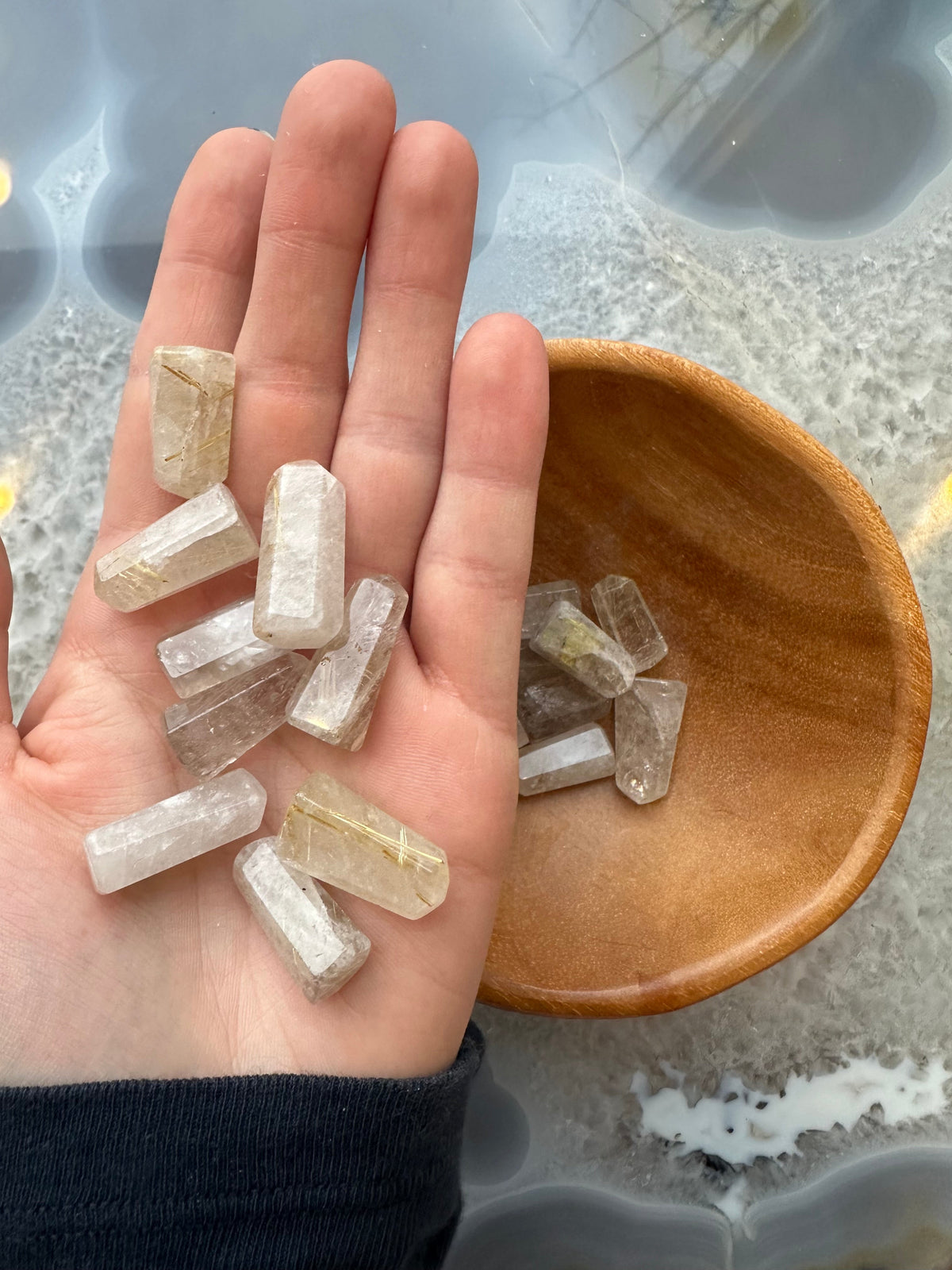 Small translucent rutilated quartz crystal points with golden needle-like inclusions held in a hand next to a wooden bowl containing more crystals on a blue-gray polished surface.