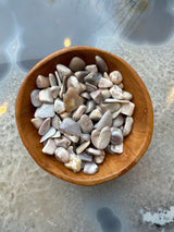 Wooden bowl filled with small, smooth tumbled stones in shades of gray, white, and beige with natural striations, placed on a translucent textured surface.