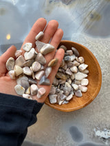 Hand holding smooth, assorted tumbled stones in cream, gray, and mauve tones above a wooden bowl filled with similar stones on a reflective surface.
