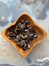 Small polished tumbled stones in varied brown, gray, and reddish hues arranged in a square wooden bowl with curved edges, set on a translucent agate slab.
