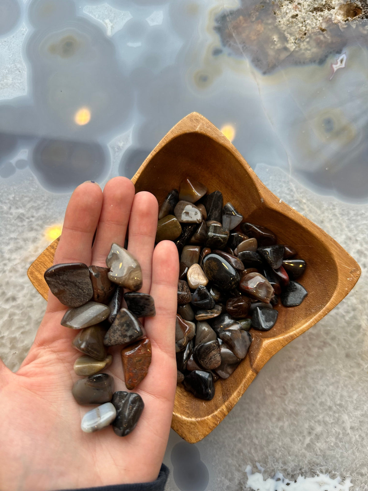 Hand holding small, polished tumbled stones in shades of brown, black, and gray next to a wooden bowl filled with similar stones, on a translucent surface.