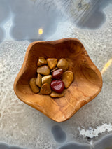 Assorted tumbled stones in warm tones of brown, tan, and red placed inside a curvy-edged wooden bowl on a translucent surface with organic patterns beneath.