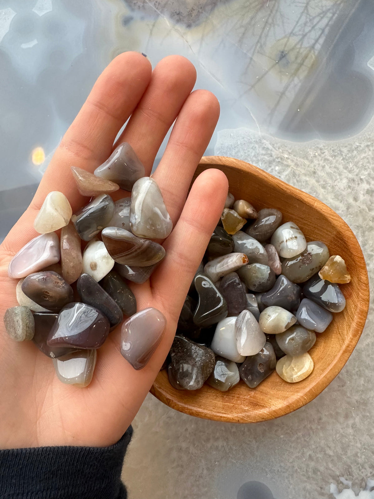 Hand holding smooth, polished tumbled stones in shades of brown, gray, and cream, with more stones in a wooden bowl on a gray surface
