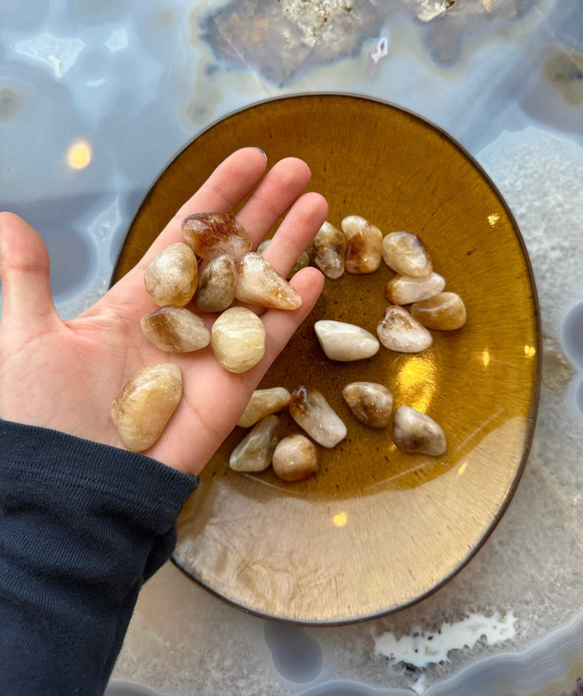 Hand holding several small tumbled citrine stones with a smooth, polished amber-brown bowl filled with more stones on a blue and gray agate surface.