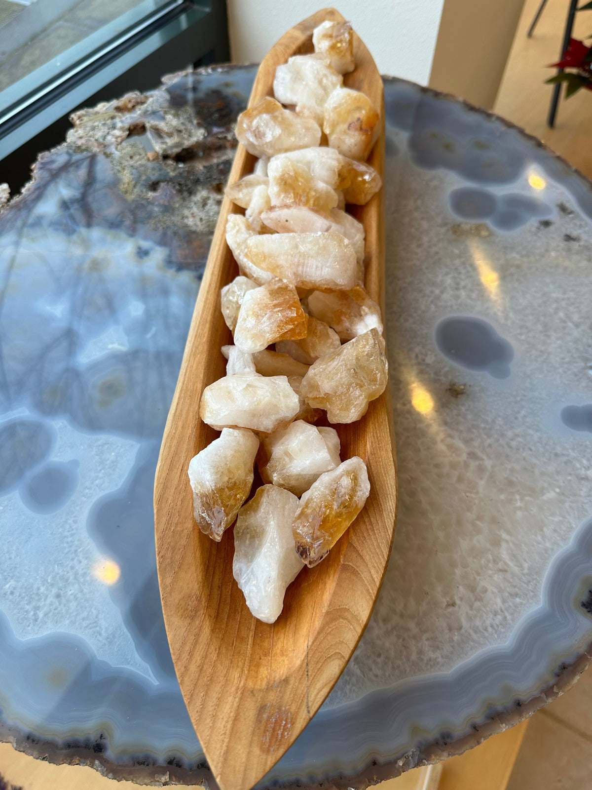 Cluster of rough citrine chunks with white and golden hues arranged in a carved wooden dish on a polished blue-gray agate slab table top.