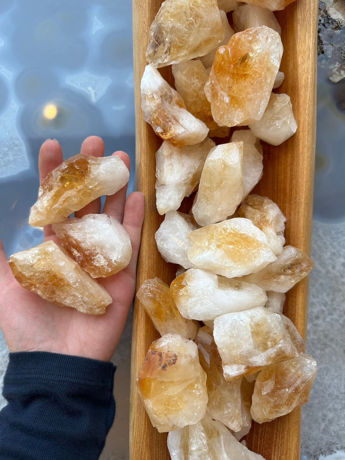 Hand holding several rough citrine chunks with amber and white hues next to a wooden tray filled with similar crystals, on a gray-blue surface.