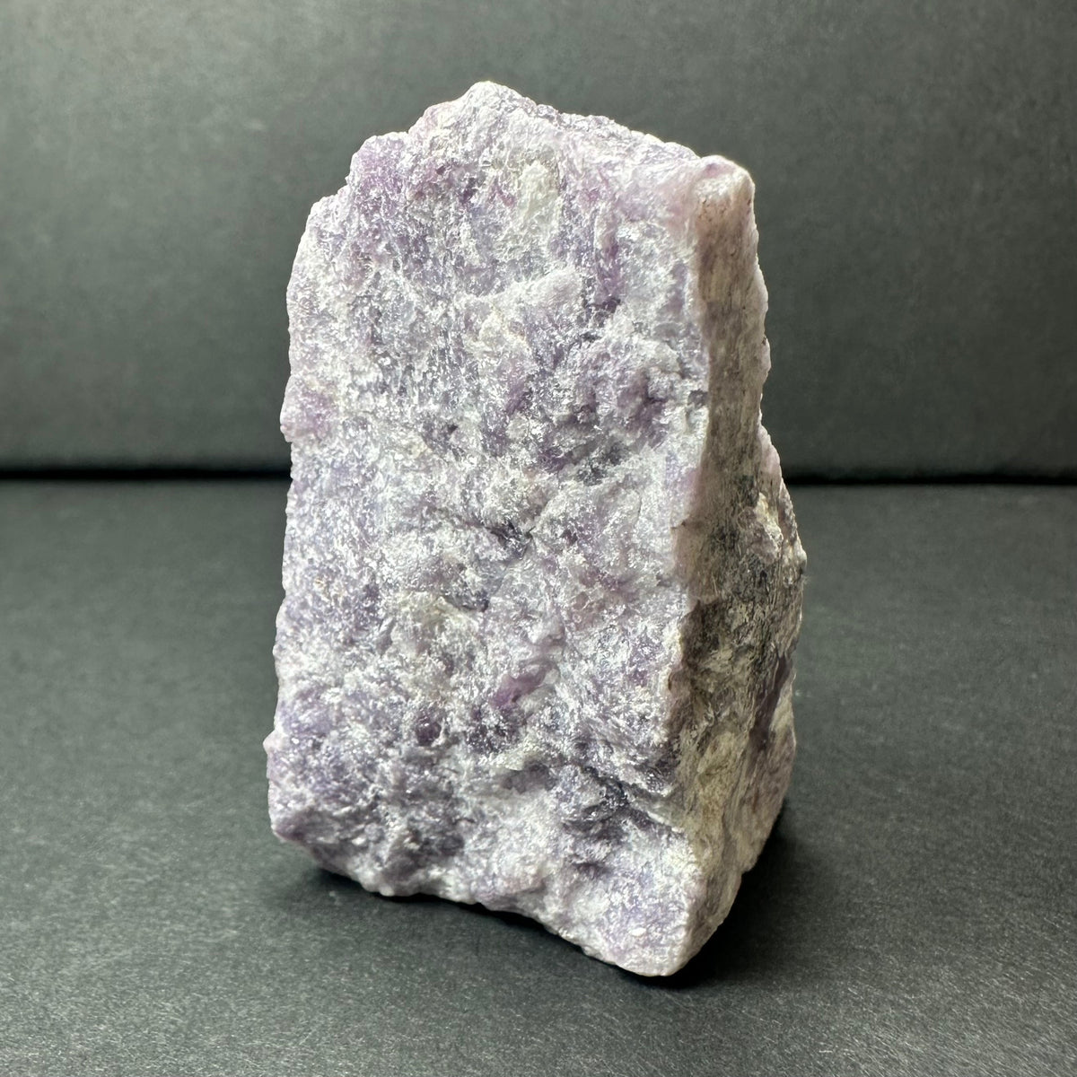 Rough purple lepidolite stone with textured, uneven surfaces and shades of lavender and white, standing upright on a dark gray background.