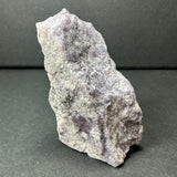 Rough, irregular lepidolite mineral displaying pale to medium purple tones with a textured, crystalline surface against a dark neutral background.