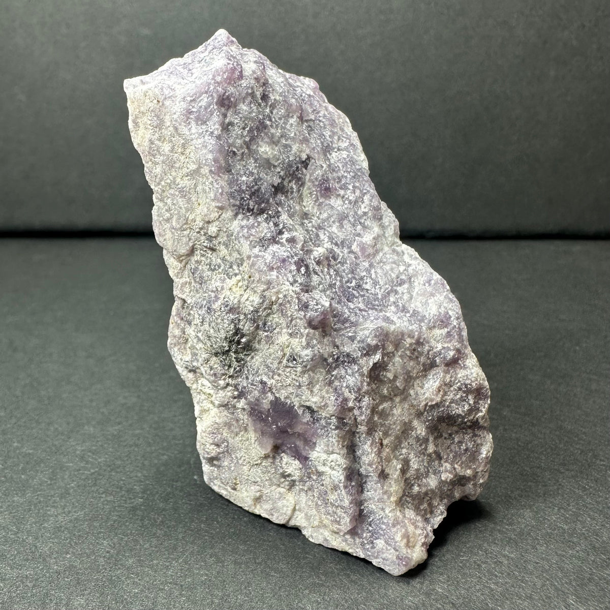 Rough, irregular lepidolite mineral displaying pale to medium purple tones with a textured, crystalline surface against a dark neutral background.
