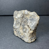 Rough labradorite stone with gray, beige, and slight iridescent tones, showing a textured, uneven surface on a dark background.