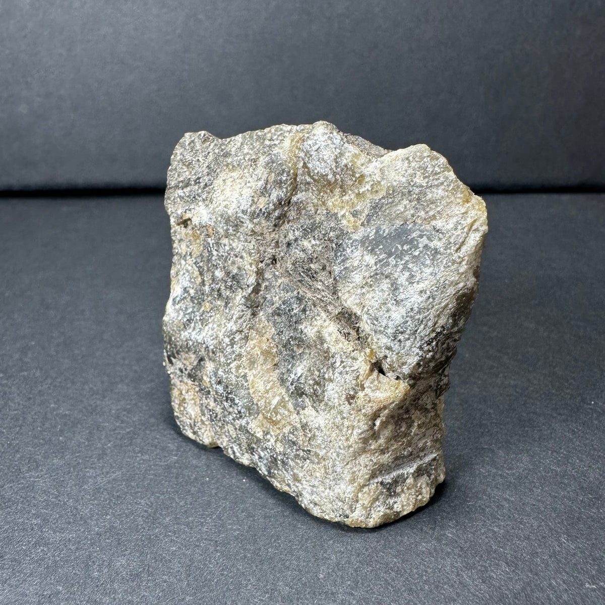 Rough labradorite stone with gray, beige, and slight iridescent tones, showing a textured, uneven surface on a dark background.