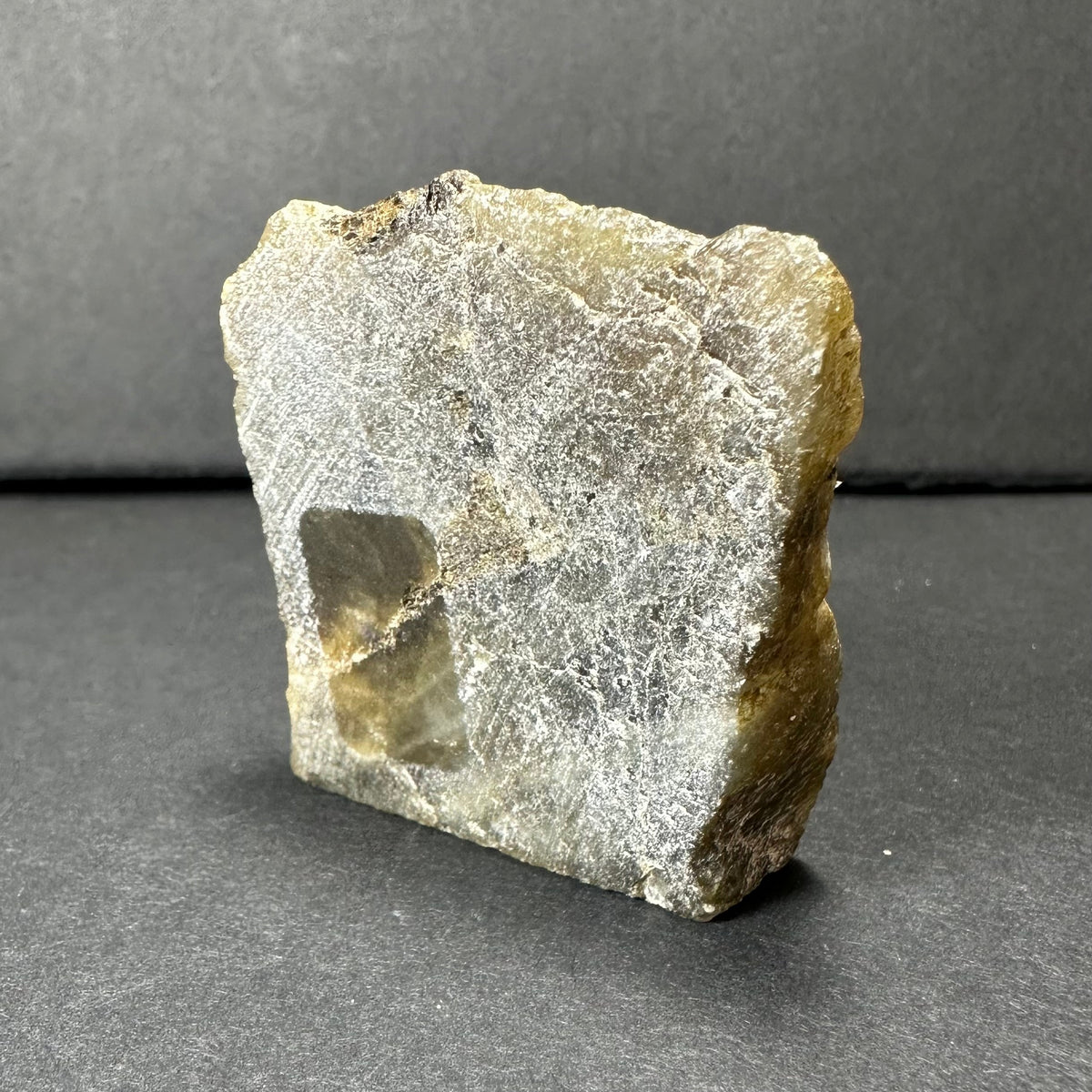 Translucent rough labradorite slab with a rectangular shape showing a pale yellow and gray surface and fibrous texture against a neutral gray background.
