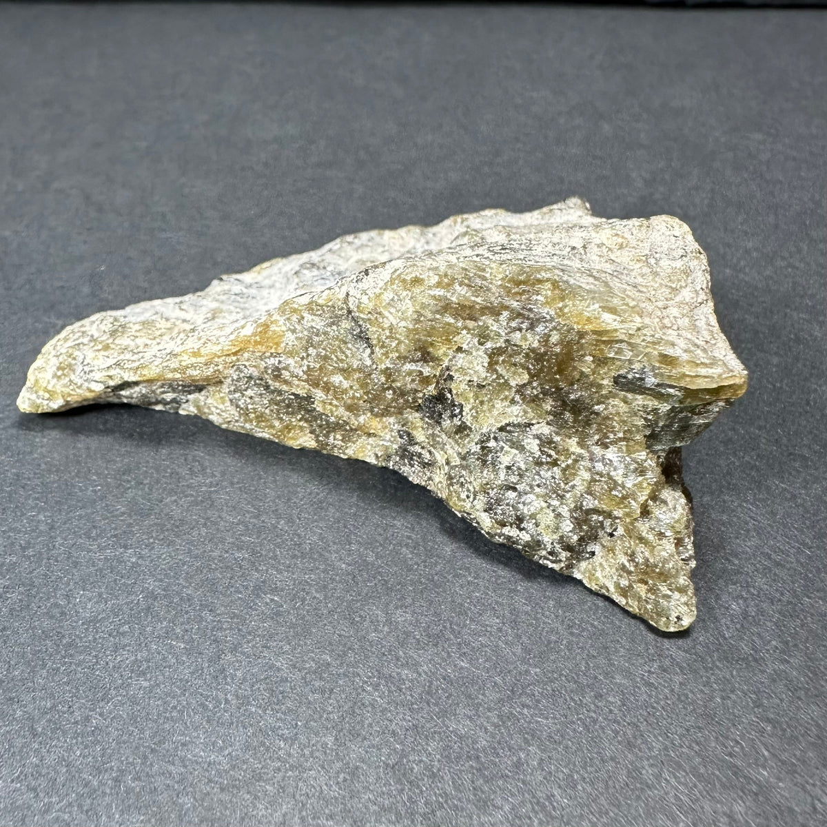 Rough labradorite stone with silvery, golden, and gray iridescent hues resting on a dark gray background, showing a textured, angular surface.