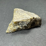 Irregularly shaped rough labradorite stone with pale yellow and gray hues resting on a dark gray surface under soft lighting.