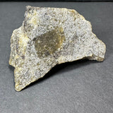 Irregularly shaped rough labradorite stone with a metallic sheen and gray, yellow, and brown tones, displayed on a dark gray surface.
