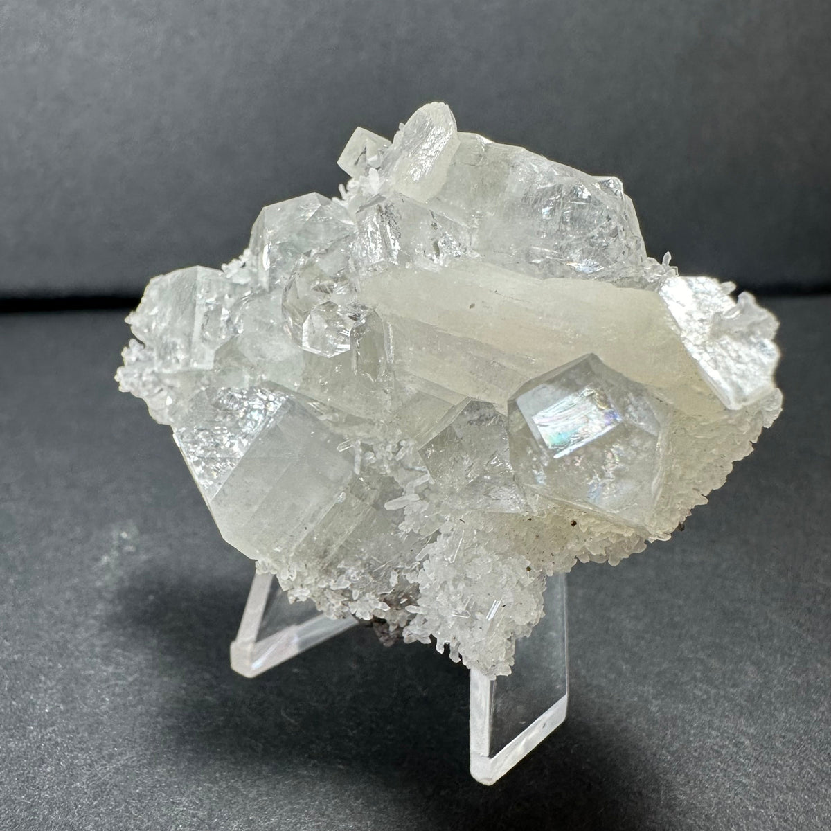 Clear apophyllite cluster with white stilbite crystals and chalcedony base displayed on a transparent stand against a dark background.