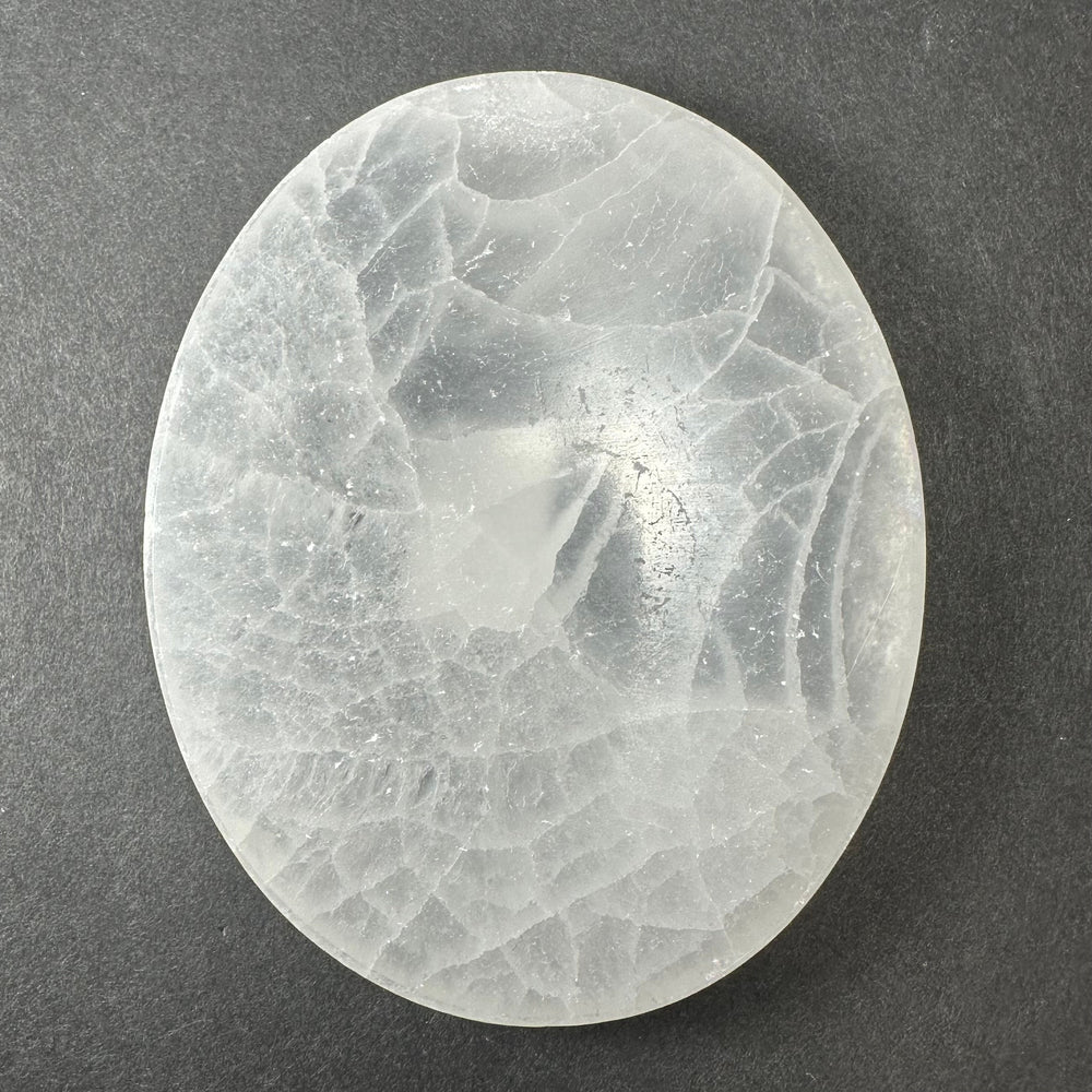 Smooth oval selenite slice with translucent white and cloudy gray patterns against a dark gray background.